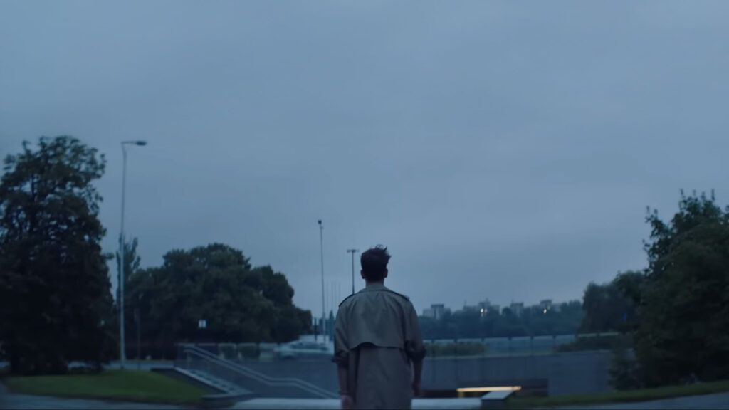 Mężczyzna w beżowym płaszczu (trenczu) idący tyłem w stronę miejskiego parku o zmierzchu, estetyka teledysku Taco Hemingwaya.