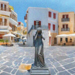 Posąg kobiety zasłaniającej twarz stoi na środku pustego placu otoczonego śródziemnomorską zabudową z białymi i pastelowymi budynkami, w słoneczny dzień.