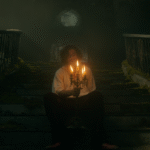 Mężczyzna siedzi na starych, porośniętych mchem schodach, trzymając w rękach kandelabr z palącymi się świecami w ciemnej, zamglonej scenerii.