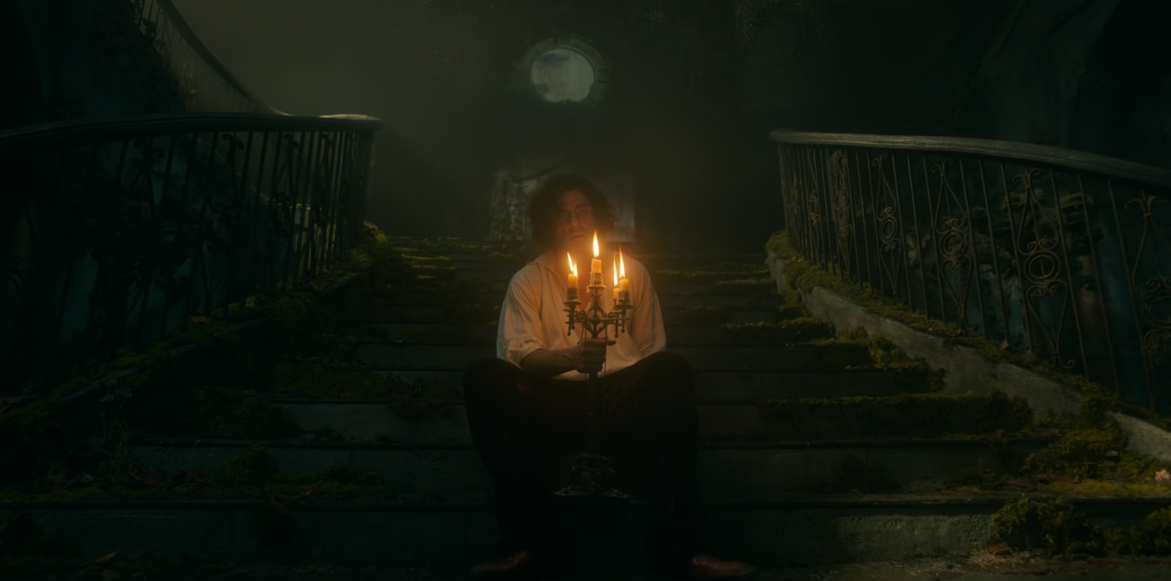 Mężczyzna siedzi na starych, porośniętych mchem schodach, trzymając w rękach kandelabr z palącymi się świecami w ciemnej, zamglonej scenerii.