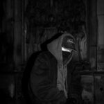 Mężczyzna w czarnej bluzie z kapturem, czapce z daszkiem i masce na tle mrocznej płaskorzeźby z postaciami – estetyka Taco Hemingway.