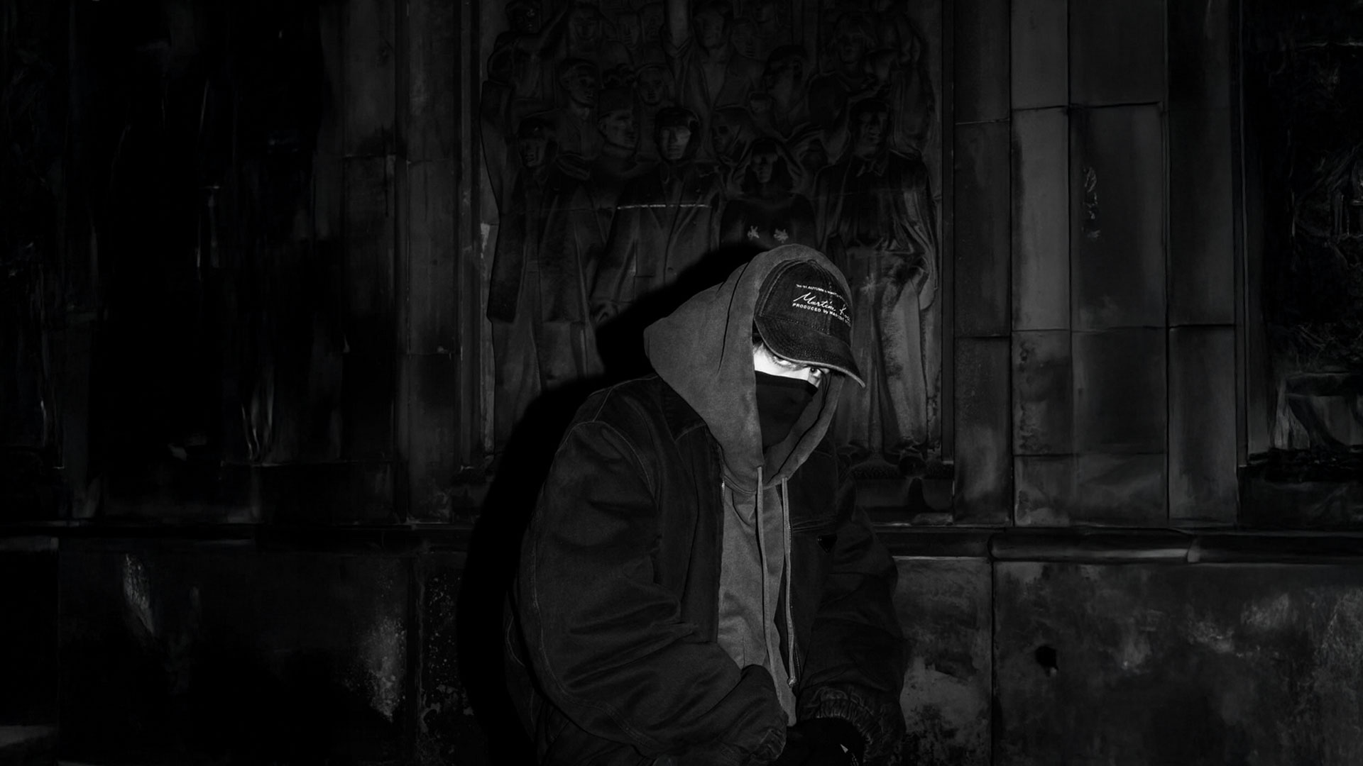 Mężczyzna w czarnej bluzie z kapturem, czapce z daszkiem i masce na tle mrocznej płaskorzeźby z postaciami – estetyka Taco Hemingway.