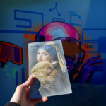 Dłoń trzyma książkę „Nadrabiam sztukę” na tle kolorowego muralu z postacią w różowym hełmie