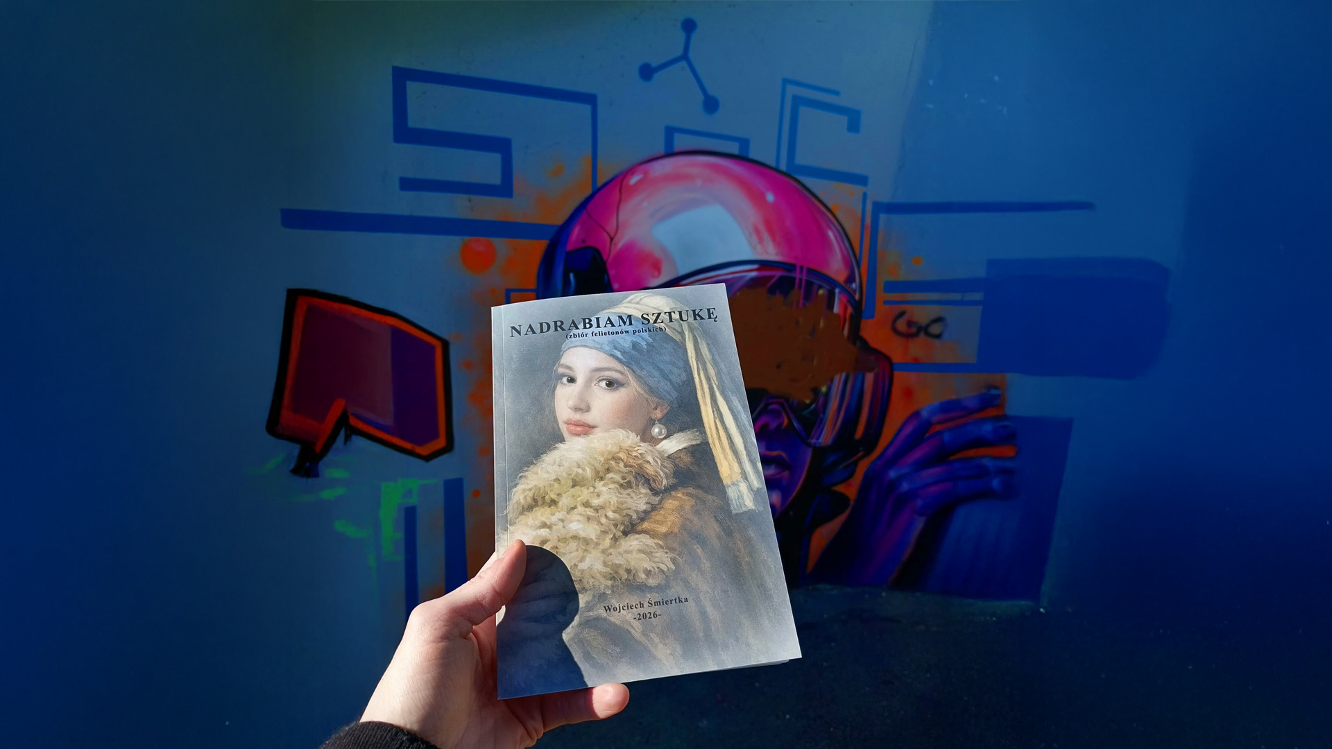 Dłoń trzyma książkę „Nadrabiam sztukę” na tle kolorowego muralu z postacią w różowym hełmie