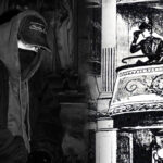 Mężczyzna w czarnej bluzie z kapturem, czapce z daszkiem i masce na tle mrocznej płaskorzeźby z postaciami – estetyka Taco Hemingway.